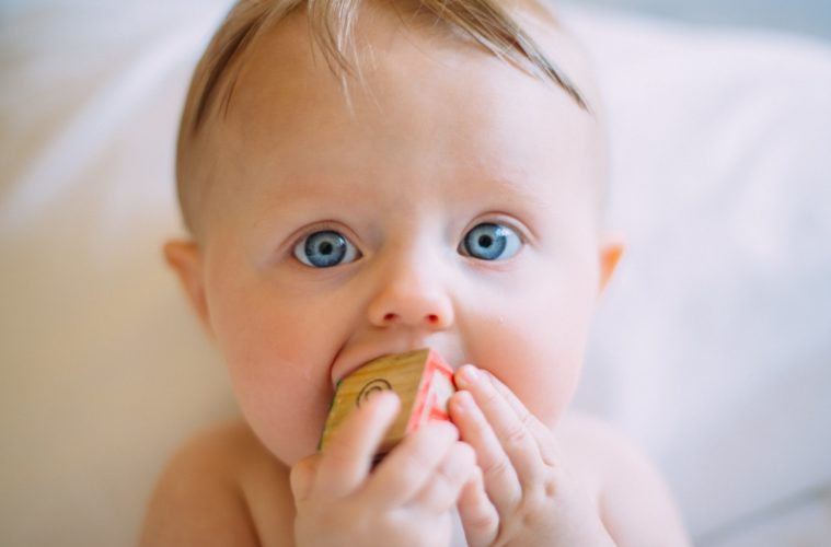 baby with blonde hair and blue eyes sucking on a block